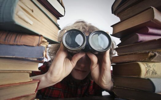 man using binoculars in between stack of books