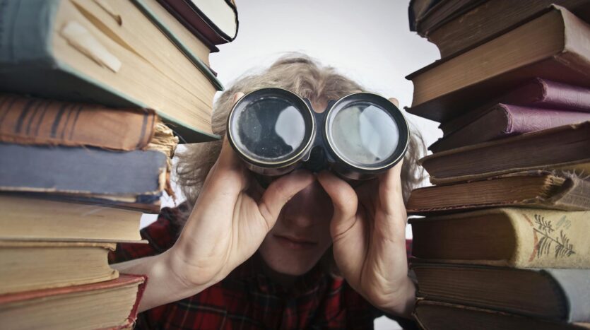 man using binoculars in between stack of books